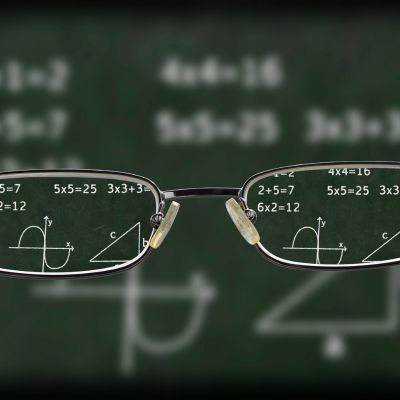 Blurry image of a chalkboard with a pair of glasses held up that you can see through clearly.