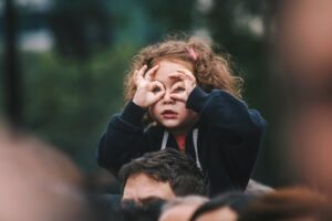 little redheaded girl in a crowed on her father’s shoulders using fingers to make circles around her eyes like glasses