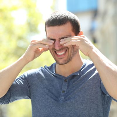 Caucasian man standing outside on a sunny day rubbing his eyes due to dry eye from seasonal allergies.