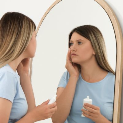 Sleep-deprived young woman covering up dark circles with concealer in front of mirror at home