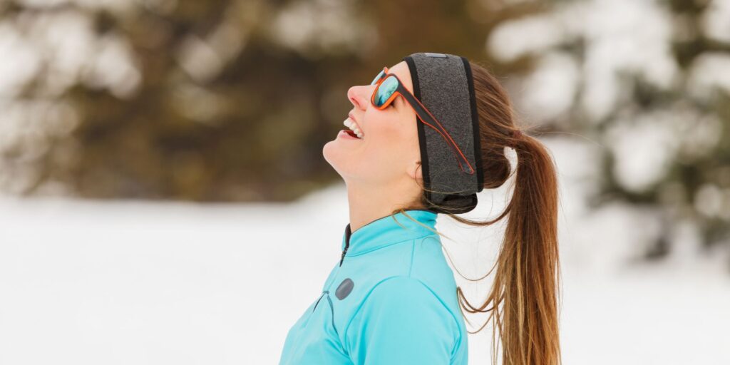 young woman in sunglasses and sports wear outside on a snowy day looking at the sky and smiling